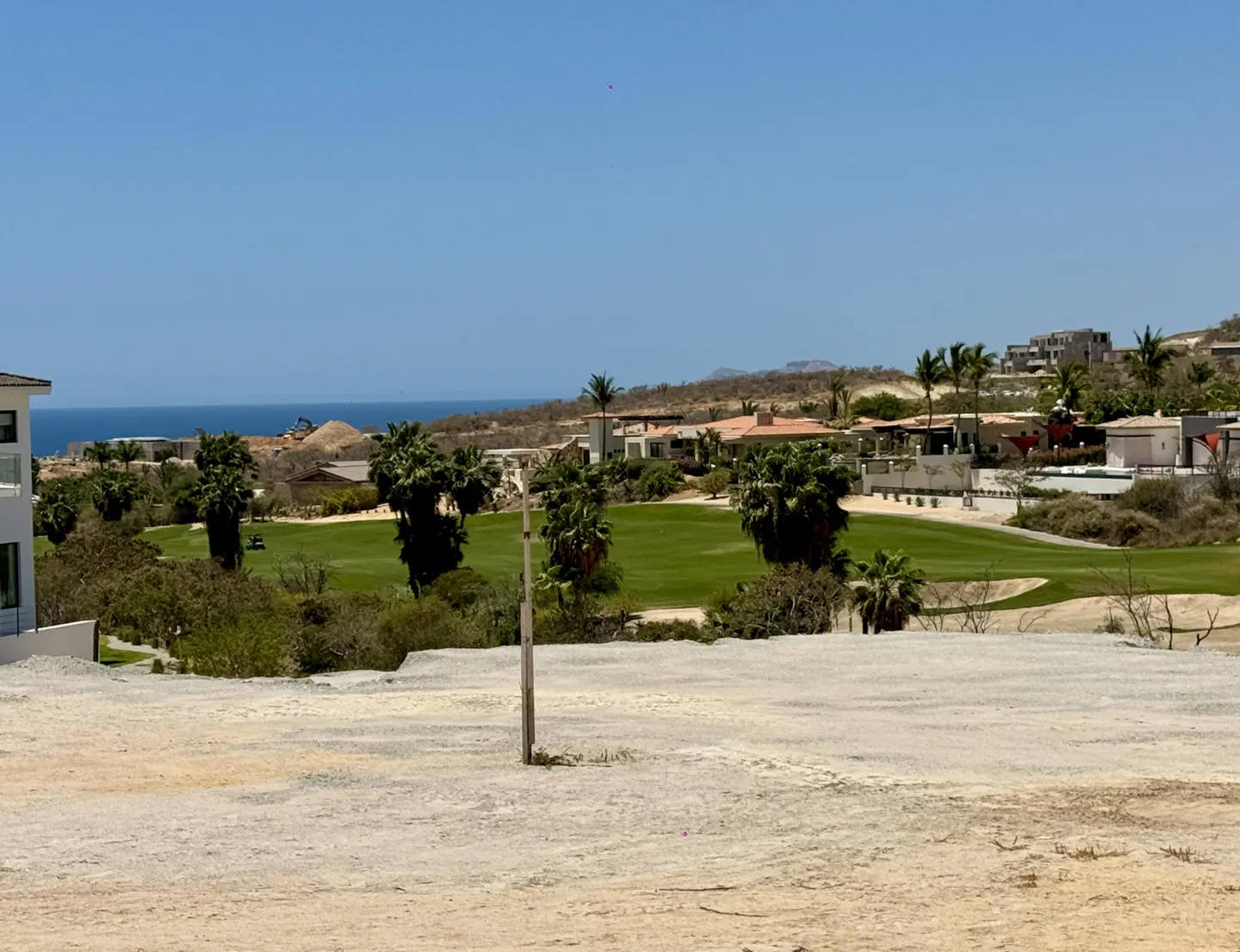 View of a vacant lot overlooking a green golf course, palm trees, and ocean under a clear blue sky.