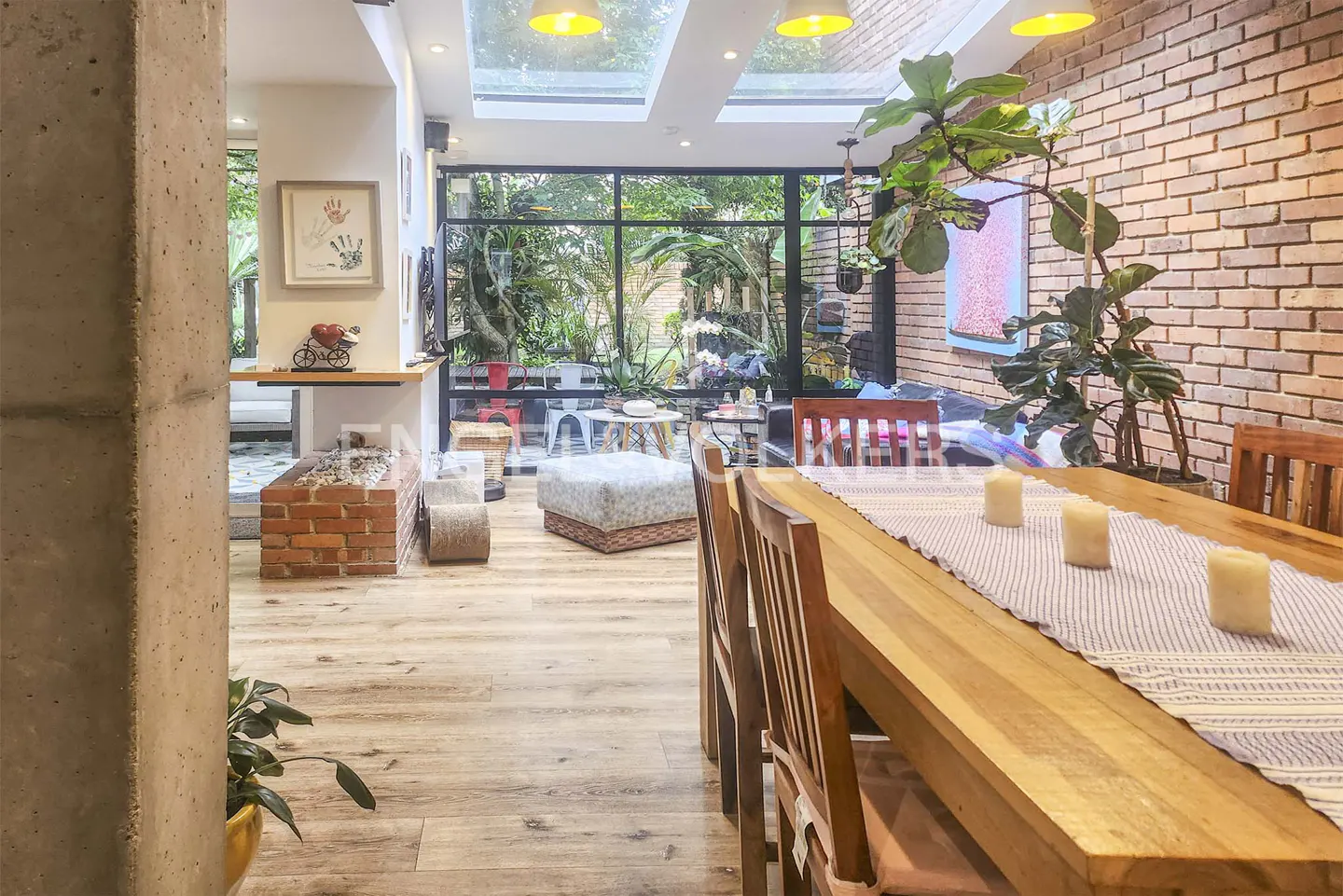 Bright dining room with a long wooden table, chairs, and candles. A brick wall and large windows overlook a lush garden.