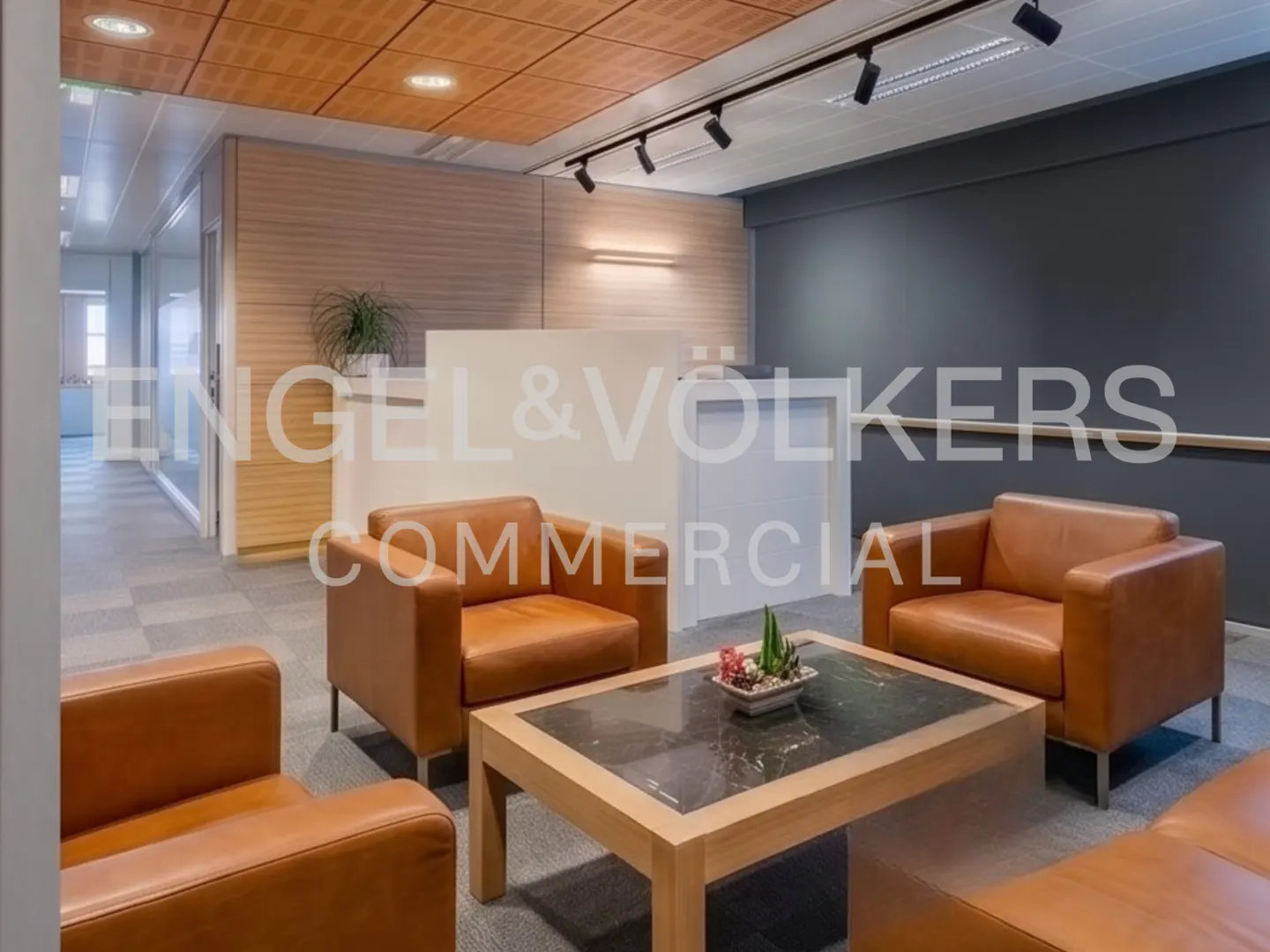 Modern office reception area with tan leather armchairs, a wooden coffee table, and a white desk against a dark wall.