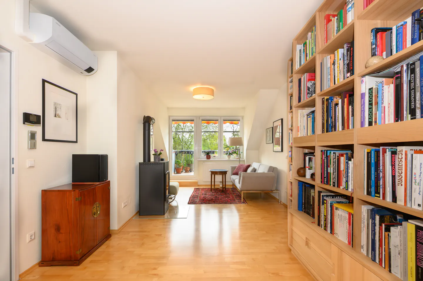 Bright living room with wood floors, a large bookcase, a black wood stove, and a gray sofa near a window.