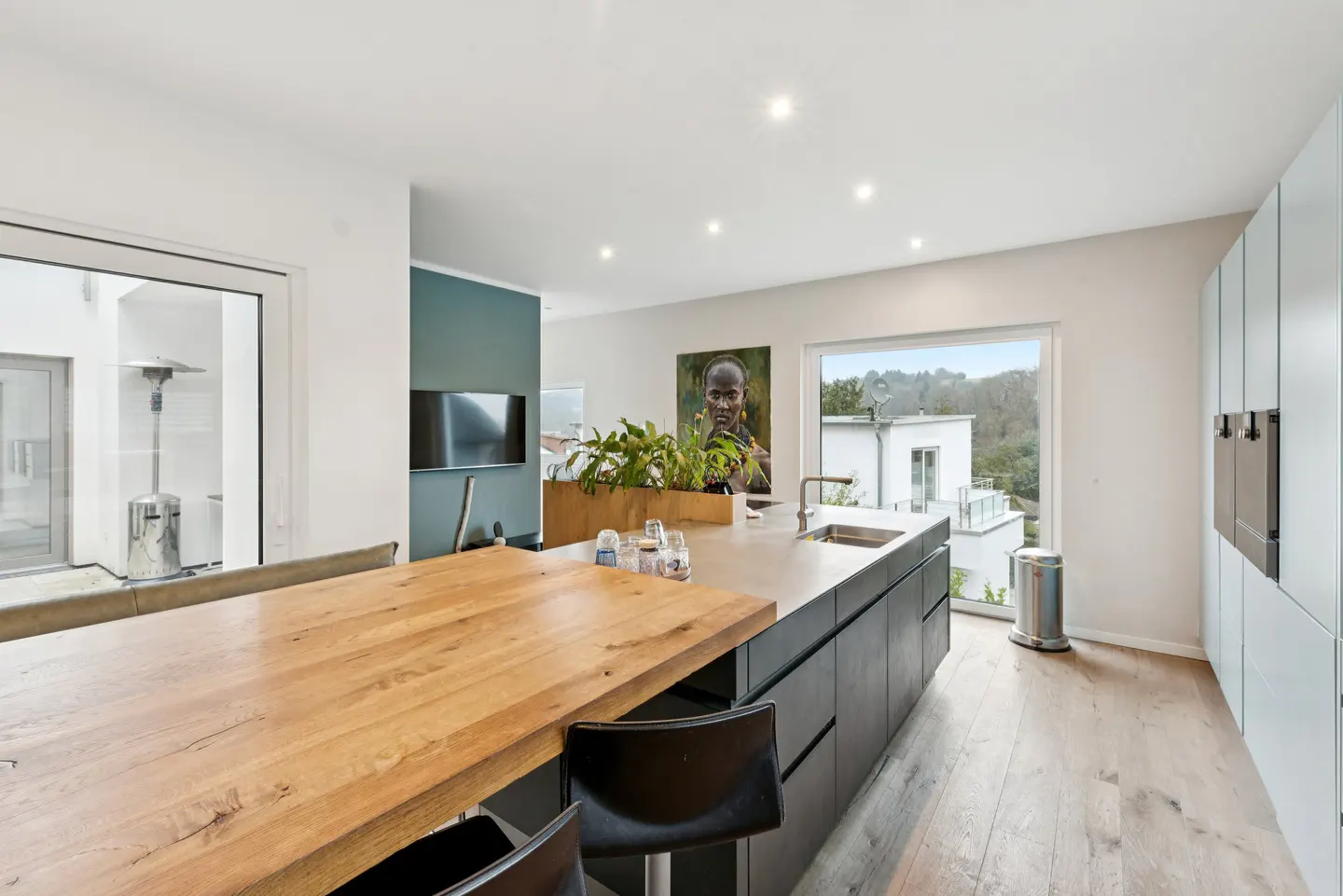A modern kitchen with a wooden table, black cabinets, and light blue walls. A large window overlooks a green landscape.
