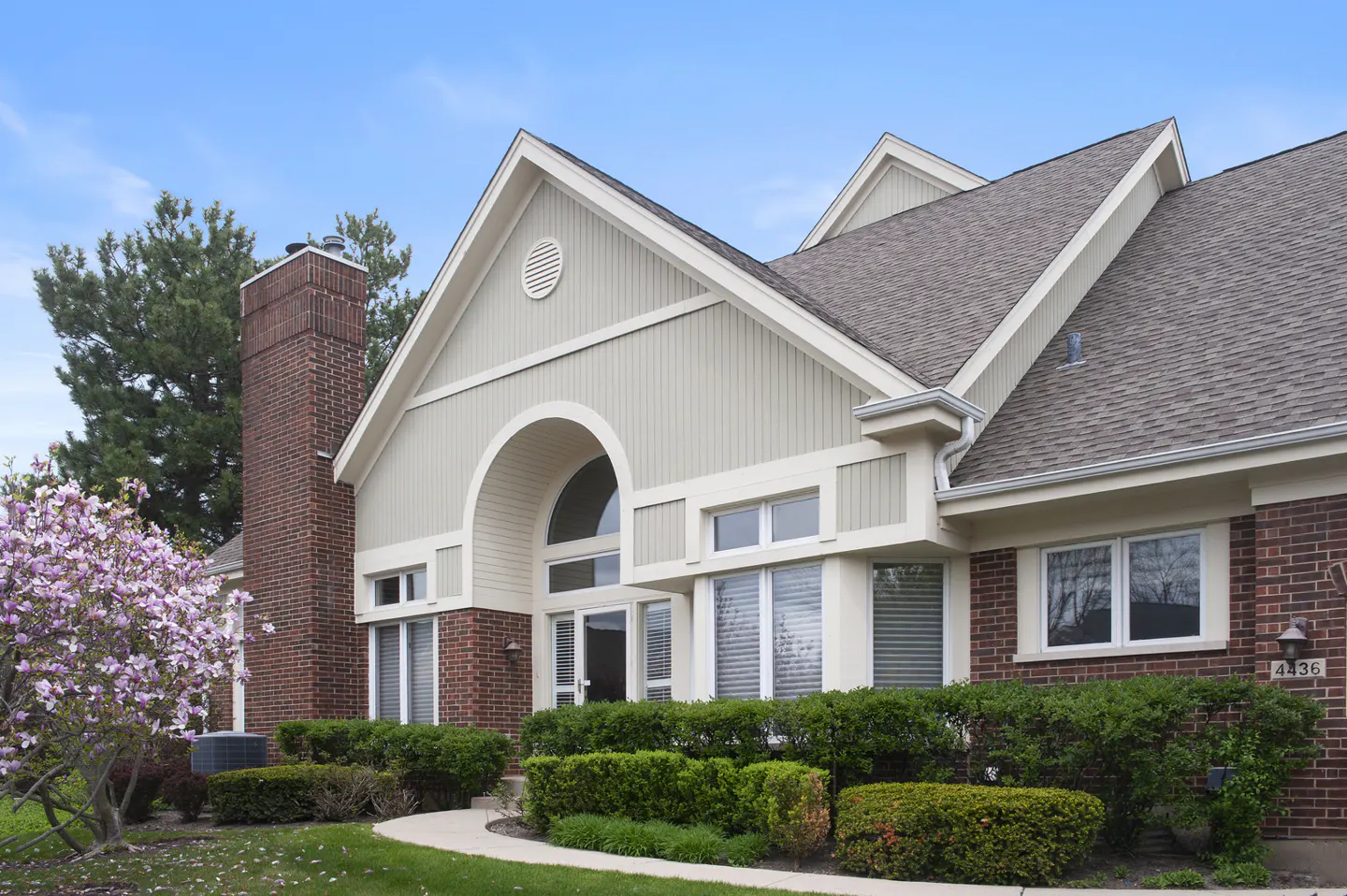 Two-story brick house with a chimney, arched window, and manicured green bushes.