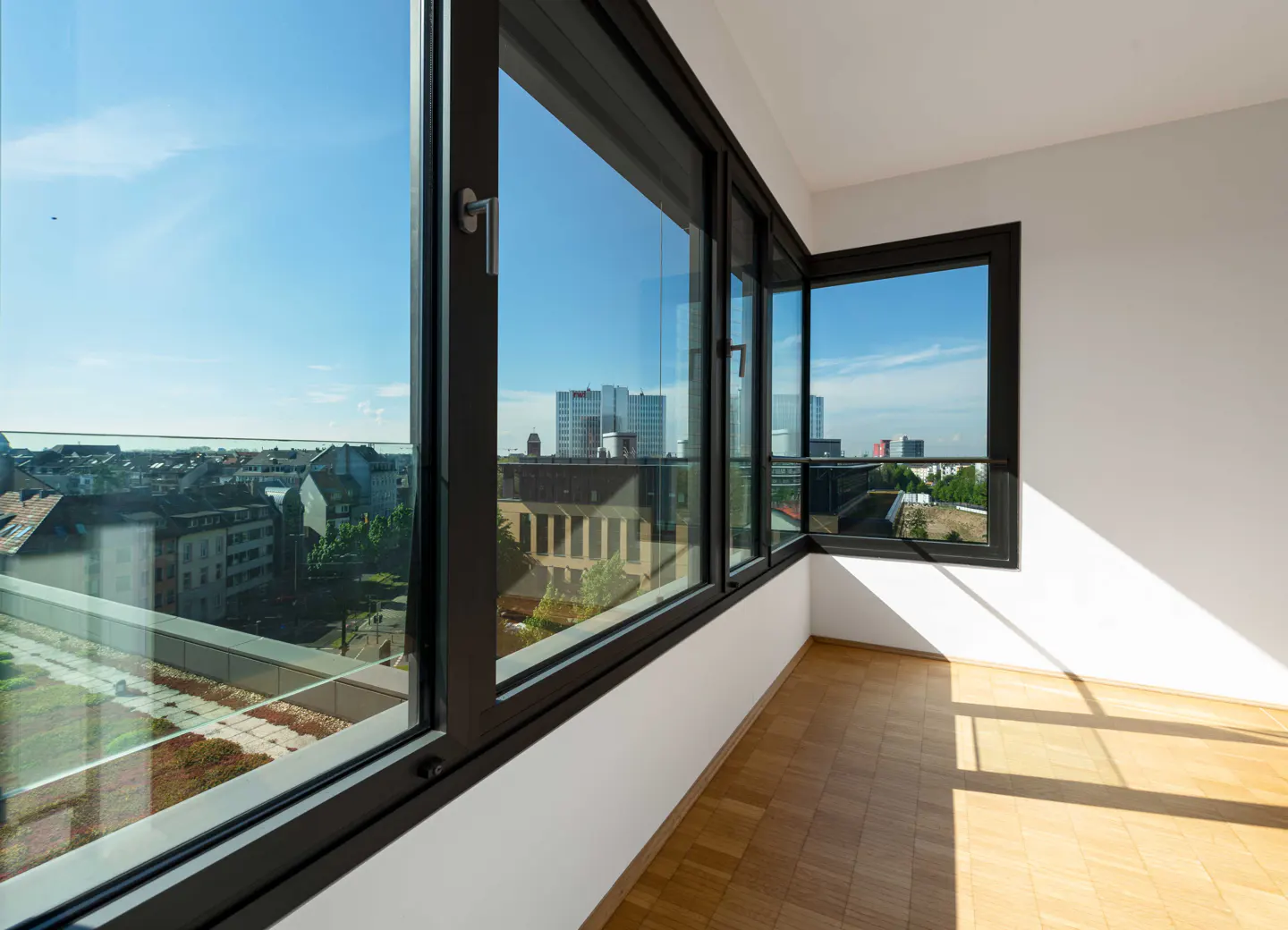 Bright room with hardwood floors and large black-framed windows showing a city view under a blue sky.