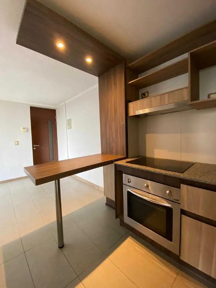 A modern kitchen with wood cabinets, stainless steel oven, and a breakfast bar extending into a living area.
