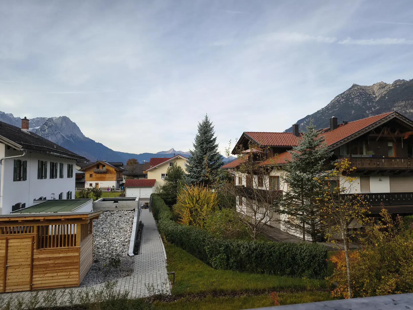 Scenic view of a European village with houses, trees, and mountains under a cloudy sky.