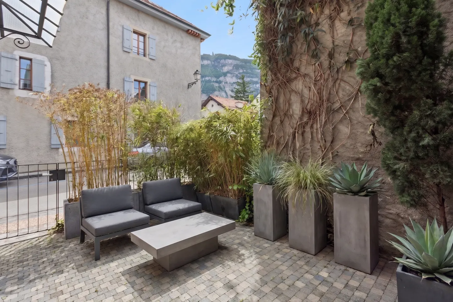 Outdoor patio with gray furniture, bamboo, and potted plants against a stone wall with climbing vines. Mountain view in the background.