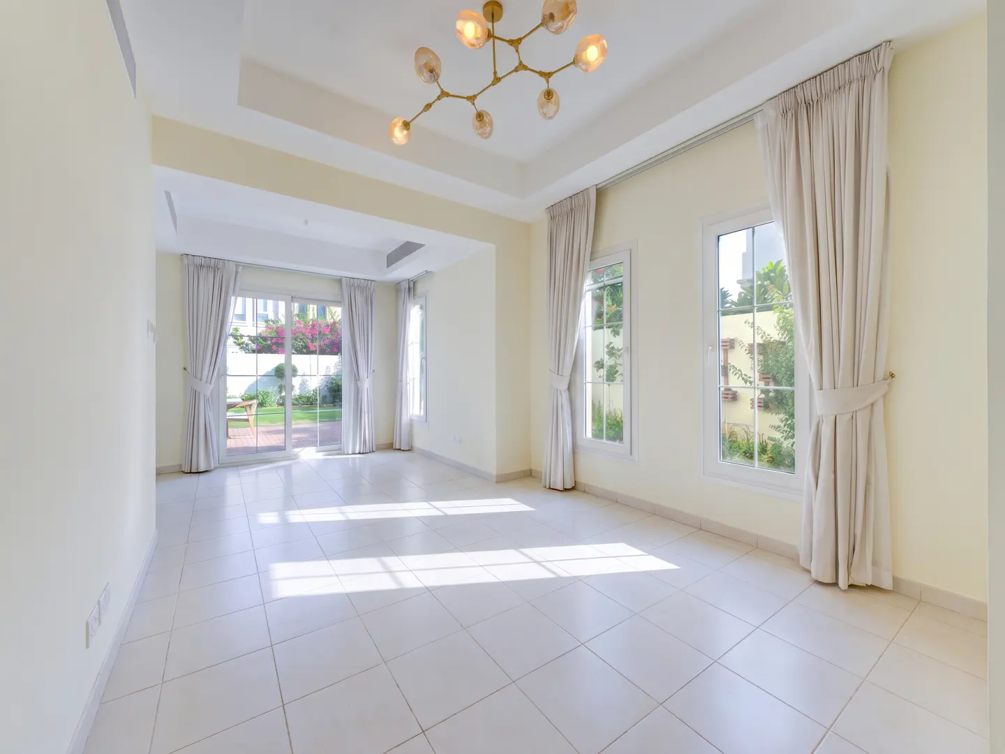 Bright, empty living room with white tiled floors, cream walls, large windows with beige curtains, and a modern chandelier.