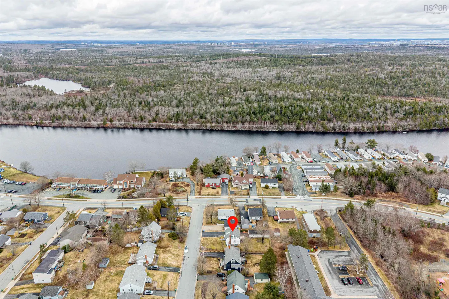 Aerial view of a residential area with a river and forest in the background. A red pin marks a property.
