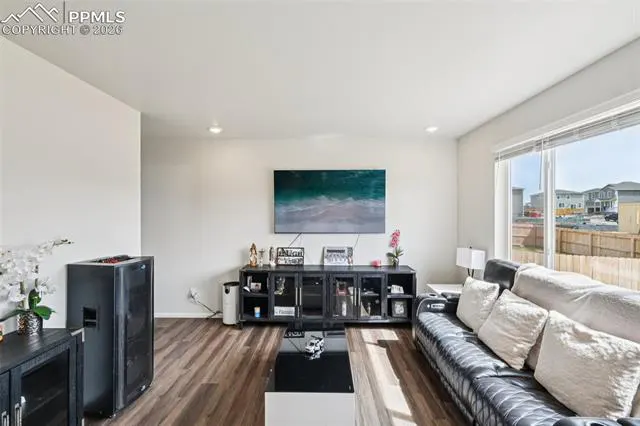 Living room with dark wood floors, white walls, and a black leather sofa. A large TV hangs above a black media console. A large window lets in natural light.
