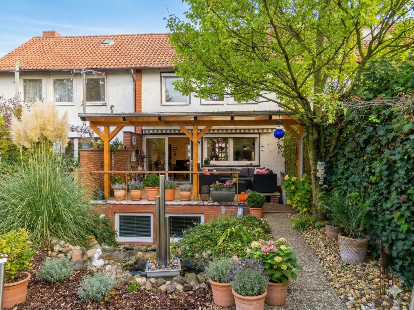 A house with a patio and garden. The patio has a wooden pergola and black furniture. The garden has a small pond and a stone path.