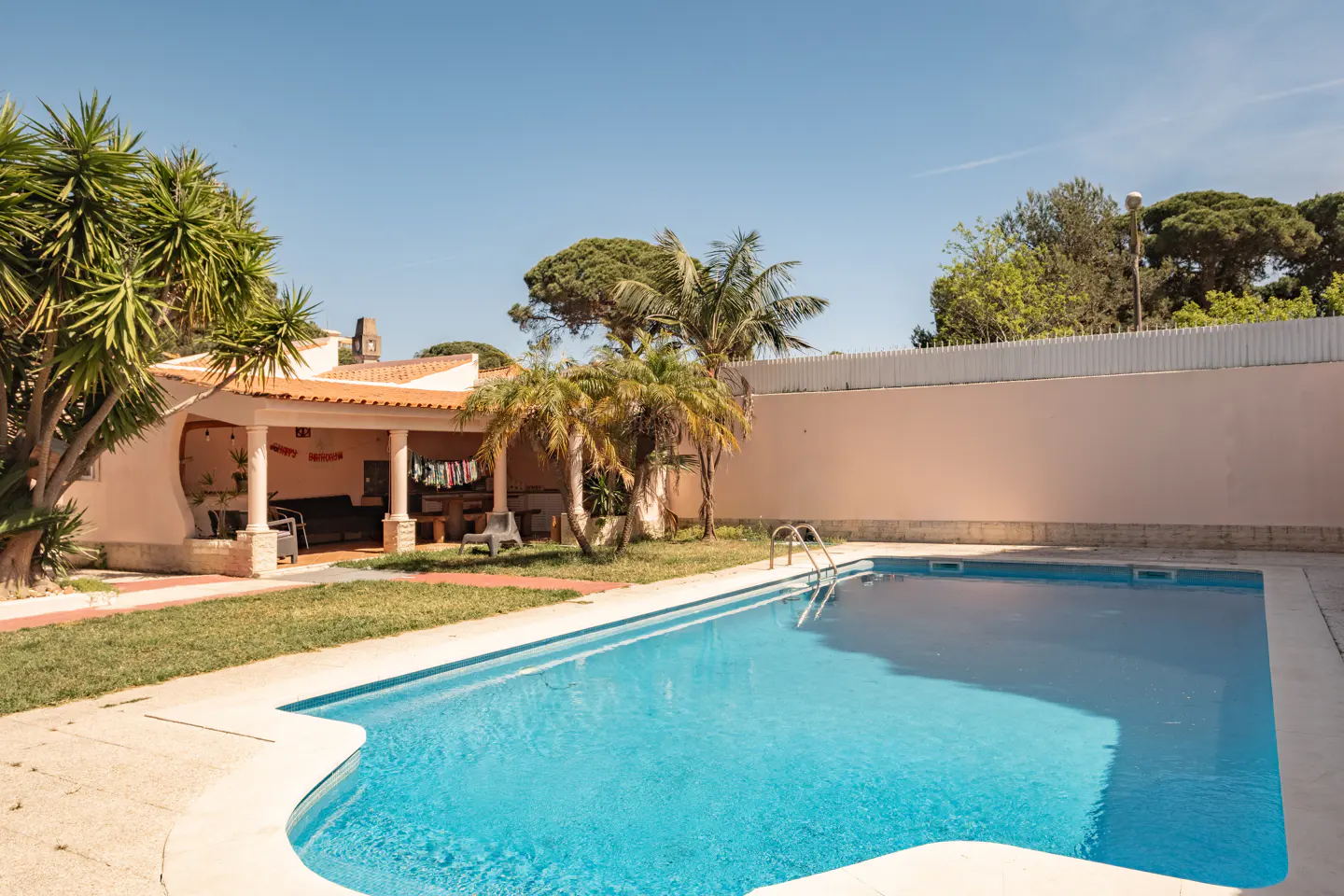 A backyard with a blue swimming pool, a covered patio, palm trees, and a light pink wall.