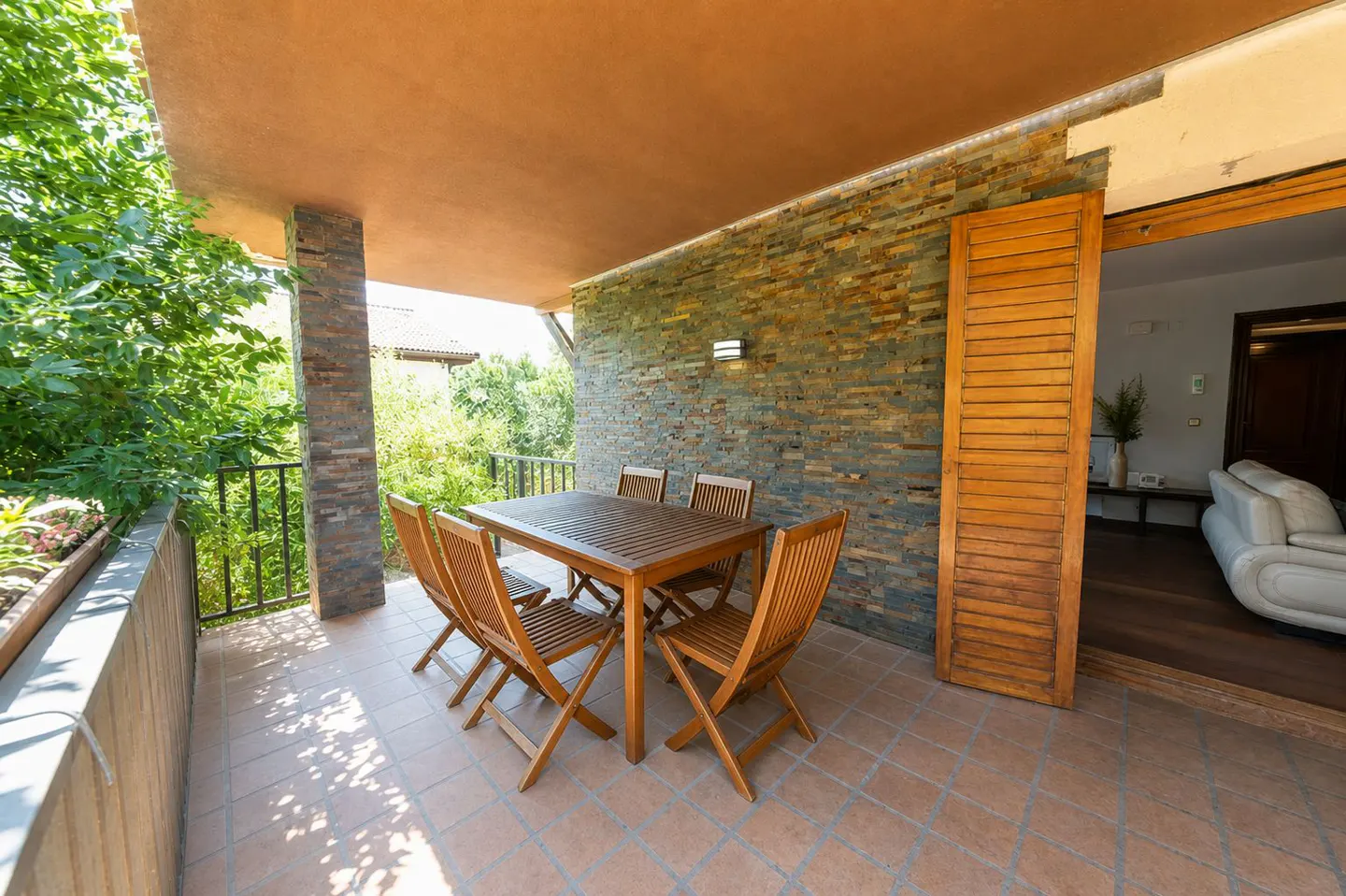 Covered patio with a wooden table and chairs. Stone wall and open shuttered door to the interior. Green trees and plants surround the patio.