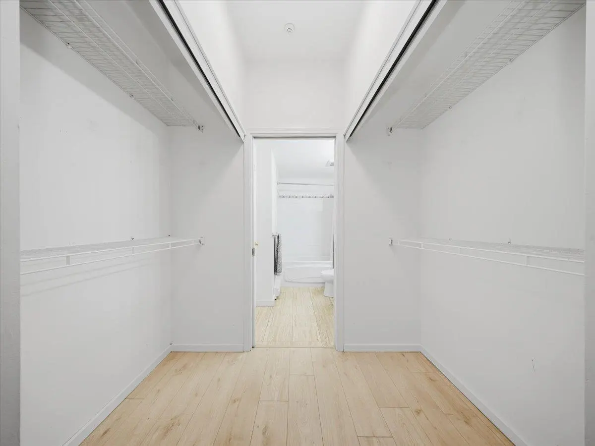 A walk-in closet with white walls, wire shelving, and light wood floors. A bathroom is visible through the open door.