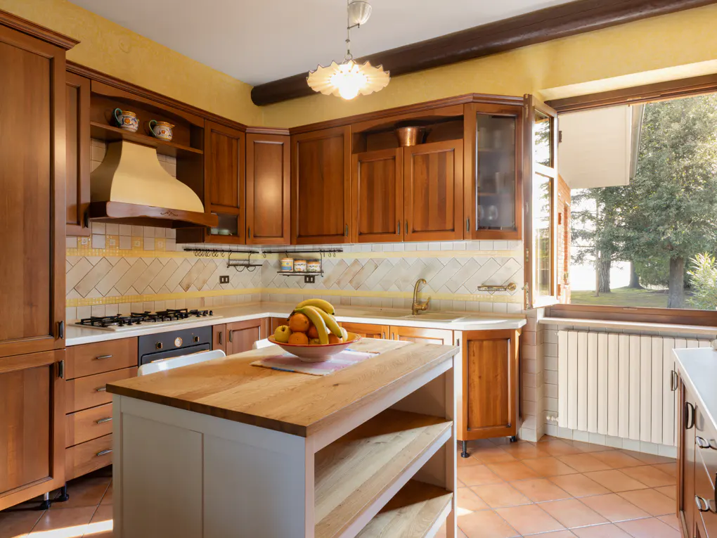 A kitchen with wood cabinets, a white island with a wood countertop, and a bowl of fruit. A window looks out onto trees.