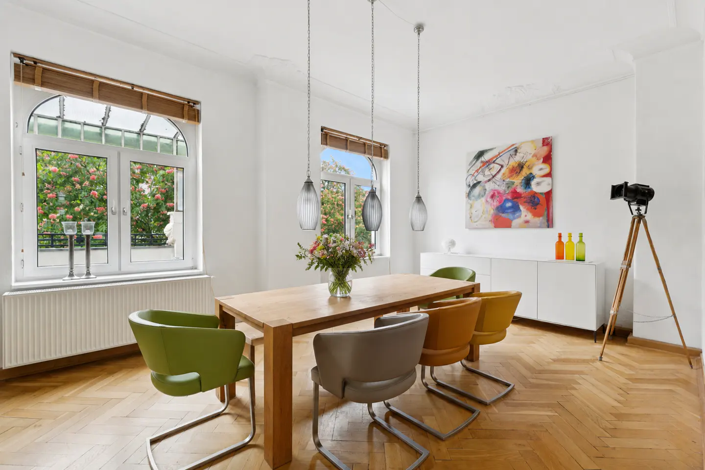 Bright dining room with wood floors, a table with colorful chairs, and a window view of pink flowers.