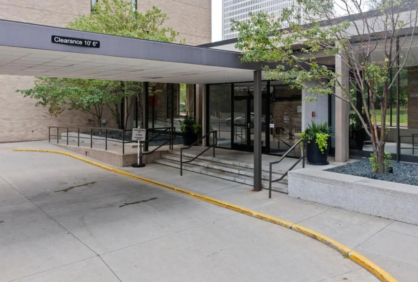 Building entrance with a 10' 6" clearance sign. Steps lead to glass doors, with trees and planters nearby. A yellow curb borders the driveway.