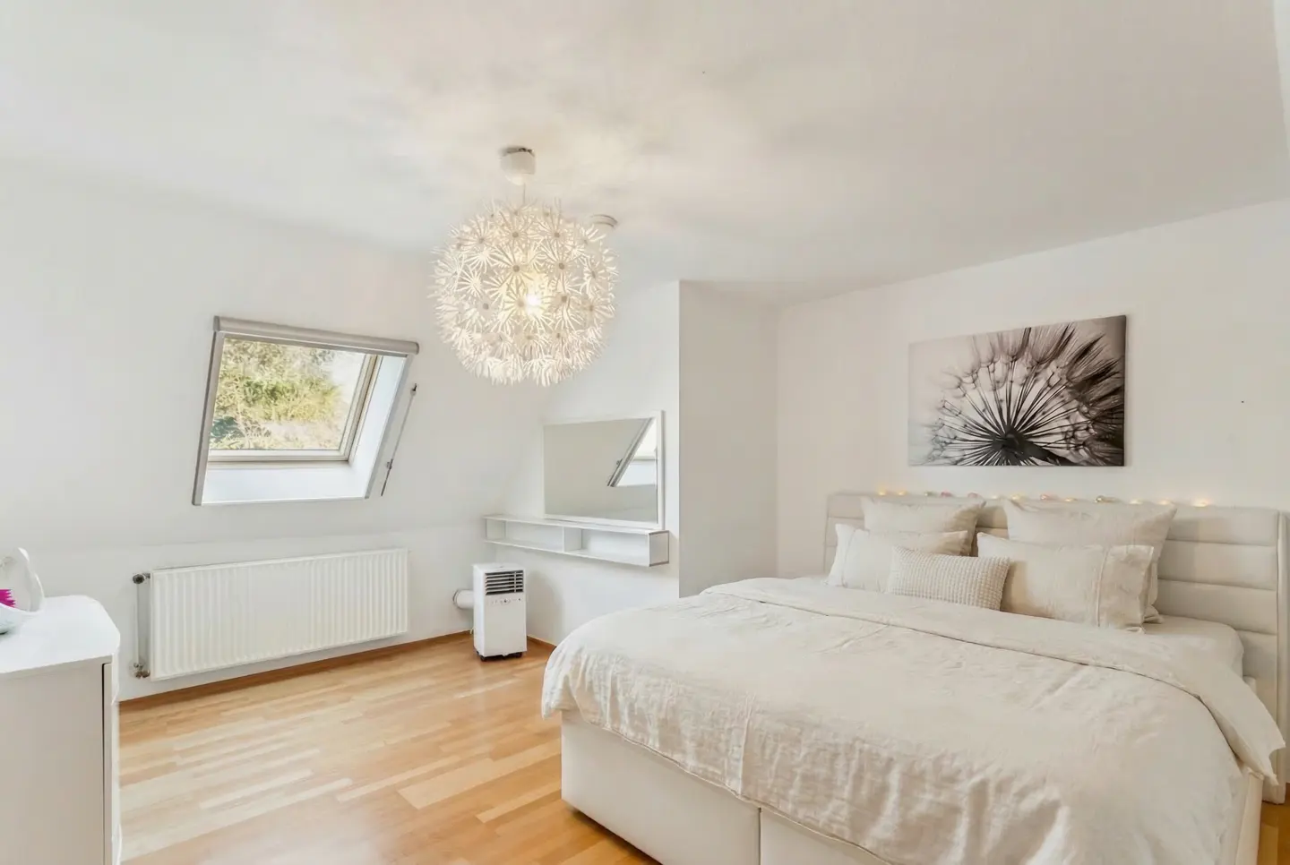 Bright bedroom with white walls, wood floor, and a skylight. A large bed with white linens is centered, with a dandelion print above. A modern light fixture hangs from the ceiling.