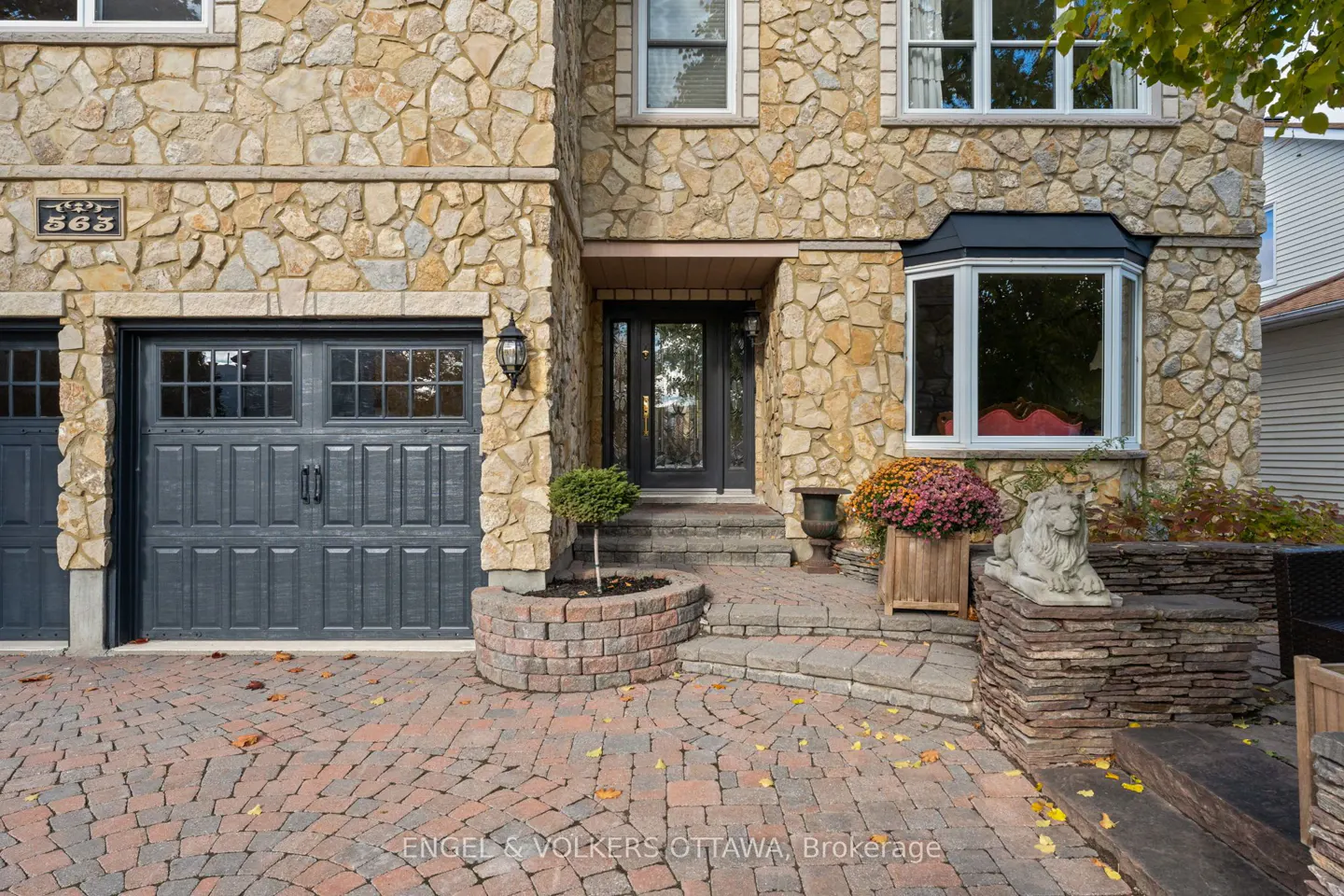 Stone house exterior with gray garage door, black front door, bay window, and brick driveway.