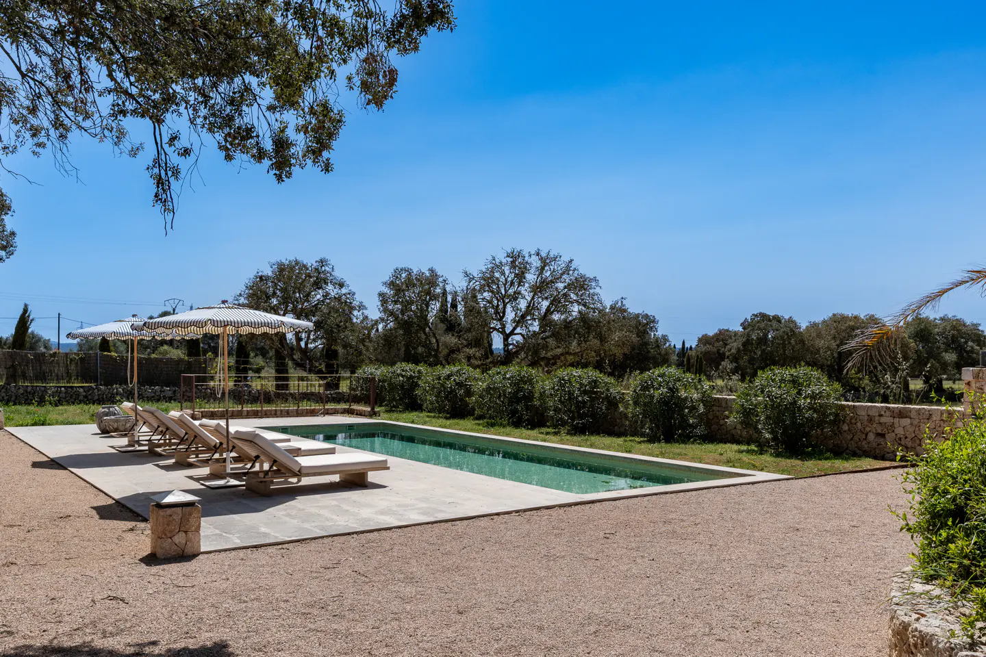 A rectangular pool with lounge chairs and striped umbrellas on a gravel patio under a clear blue sky.