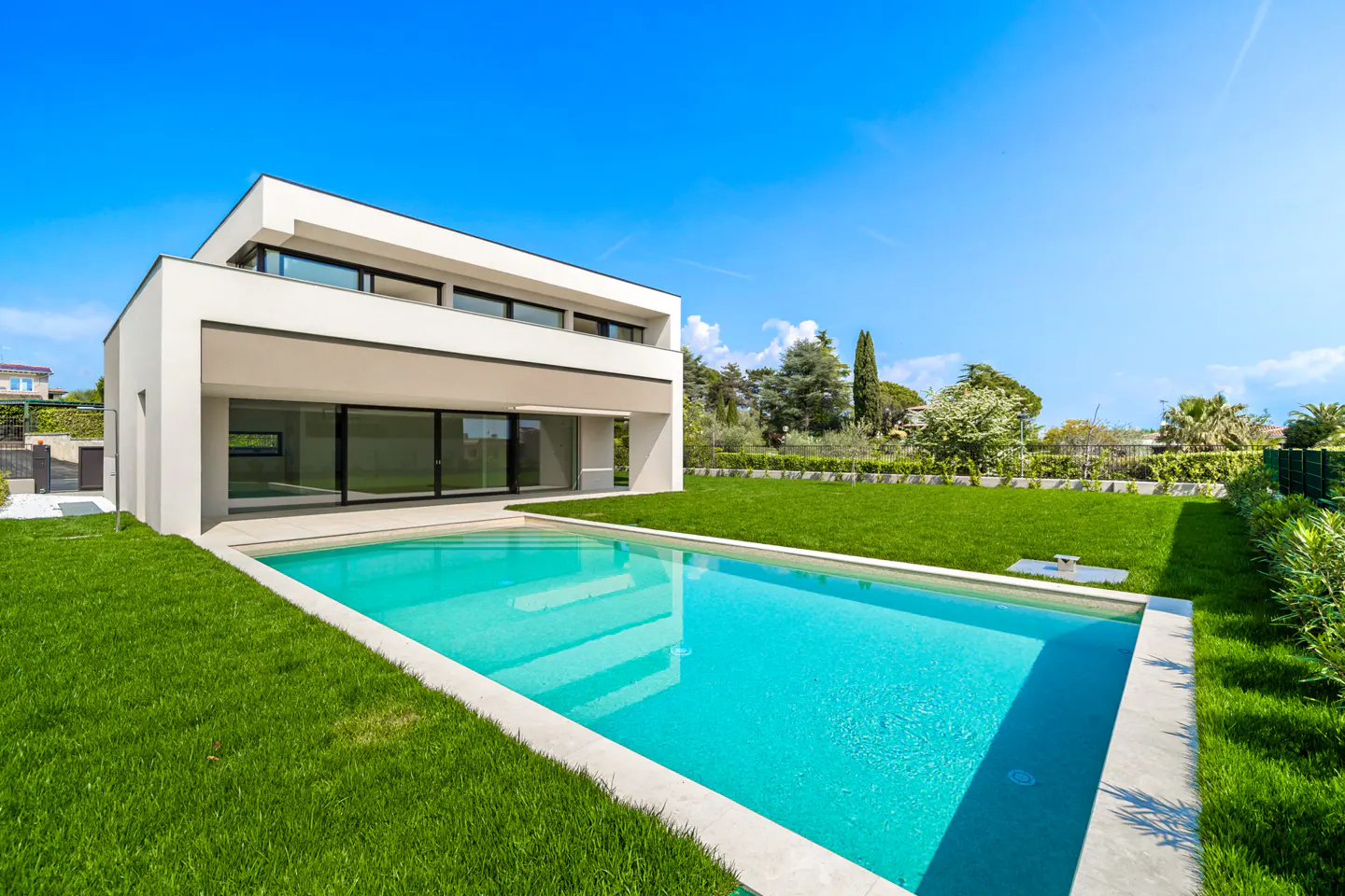 Modern two-story home with a turquoise pool, green lawn, and blue sky. The house has large windows and a flat roof.