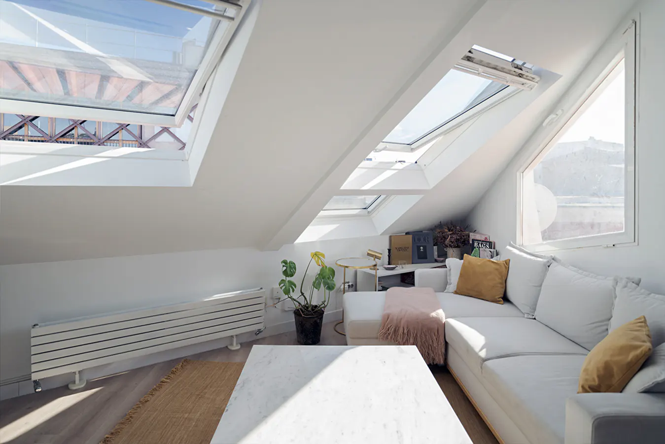 Bright, white attic living room with skylights, a white sectional sofa with yellow pillows, and a marble coffee table.