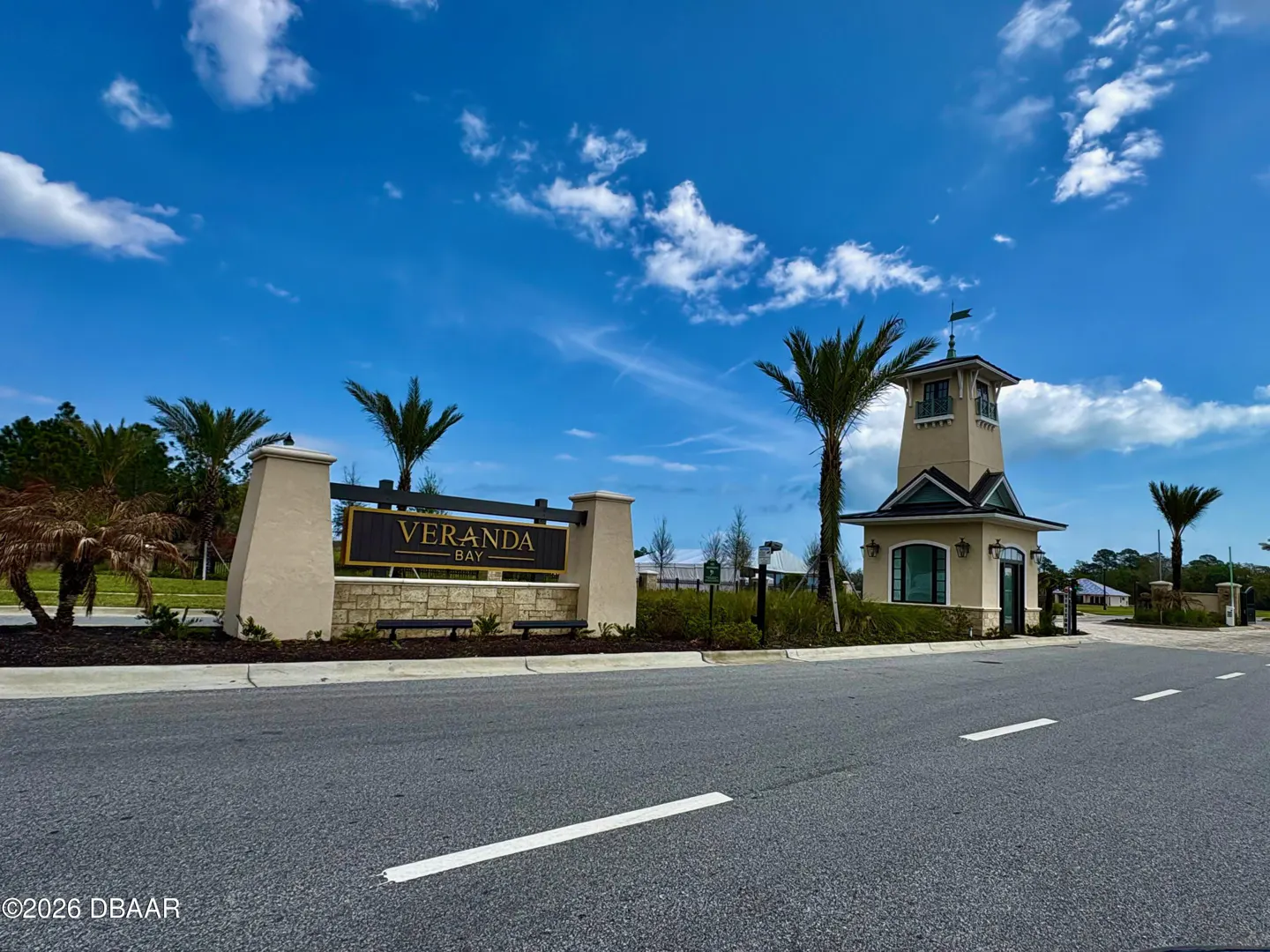 Entrance to Veranda Bay with a beige guard house and sign. Palm trees and a blue sky with clouds are in the background.