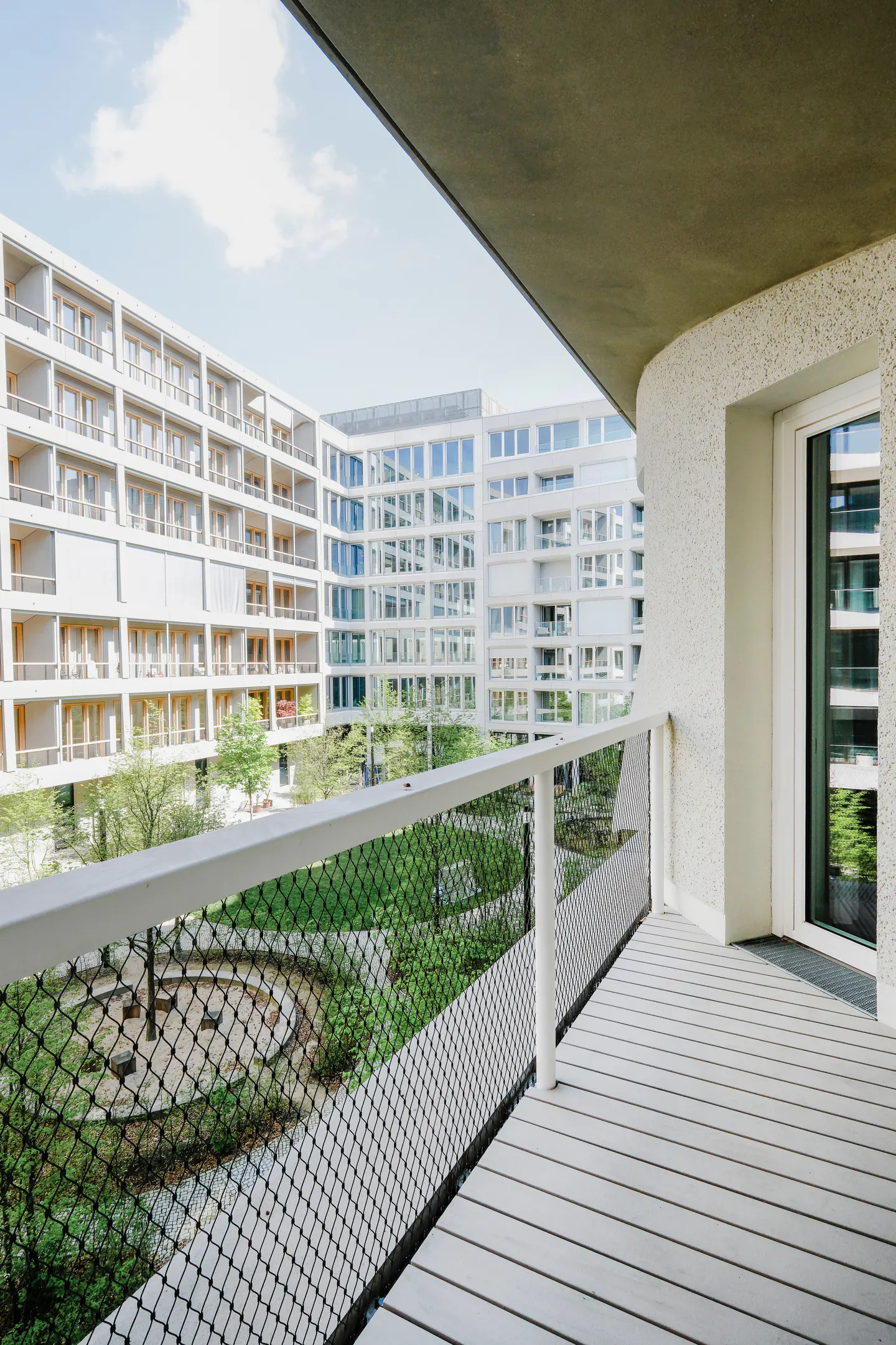 View from a white balcony with a black net railing overlooking a green courtyard and modern white apartment buildings.
