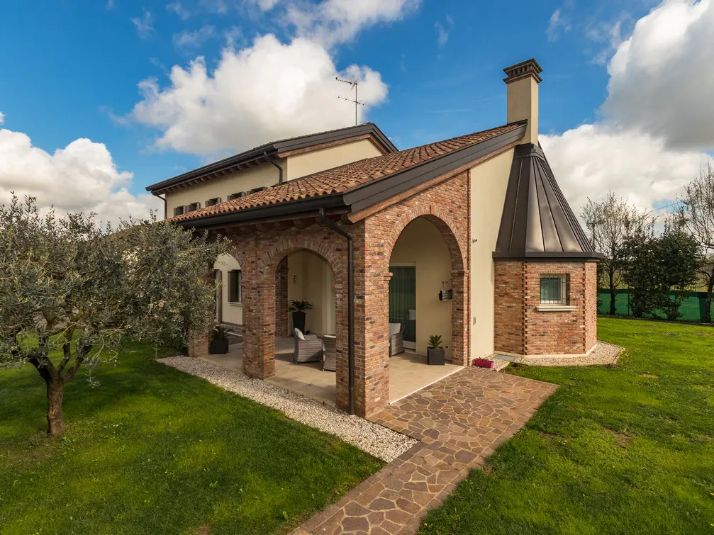 Exterior view of a beige house with brick arches, a stone walkway, and a green lawn under a blue sky with white clouds.