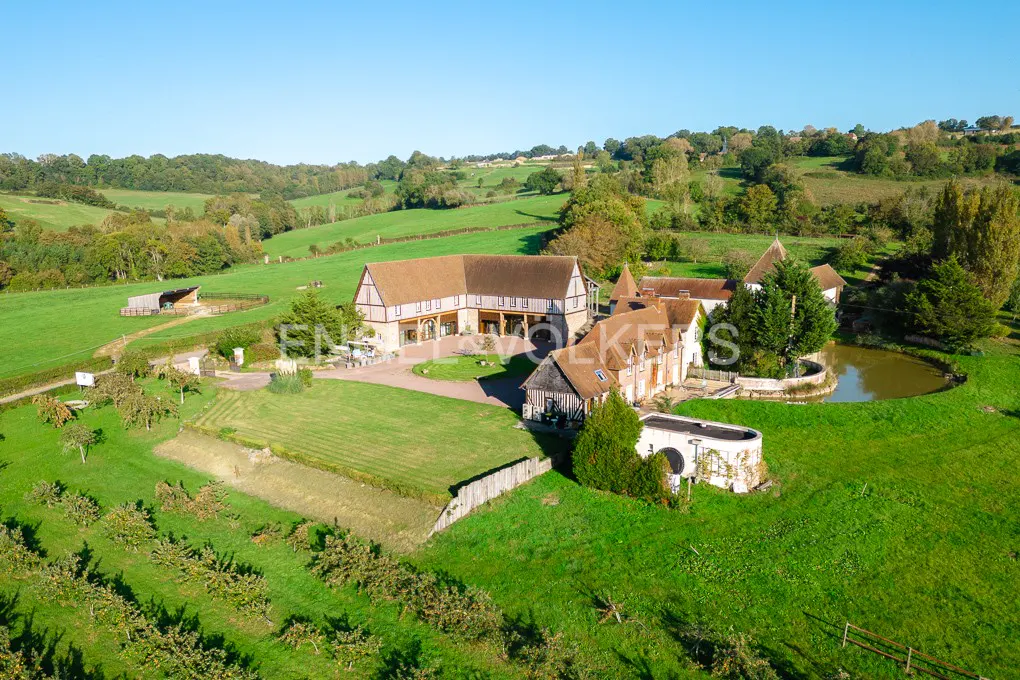 Aerial view of a large, tan-roofed house with timber framing, surrounded by green fields, trees, and a pond on a sunny day.