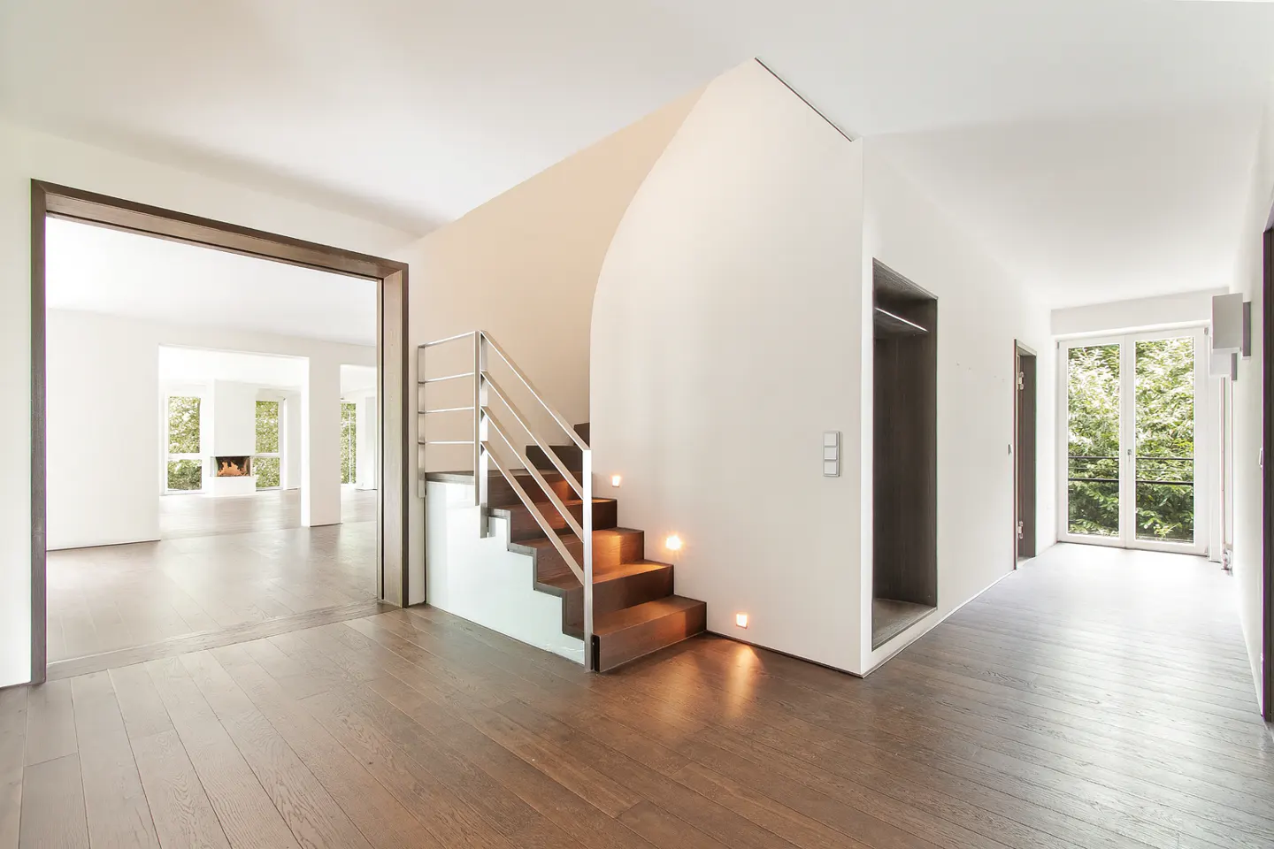 Bright, modern home interior with hardwood floors. A staircase with metal railings and an elevator are visible. A living room with a fireplace is in the background.