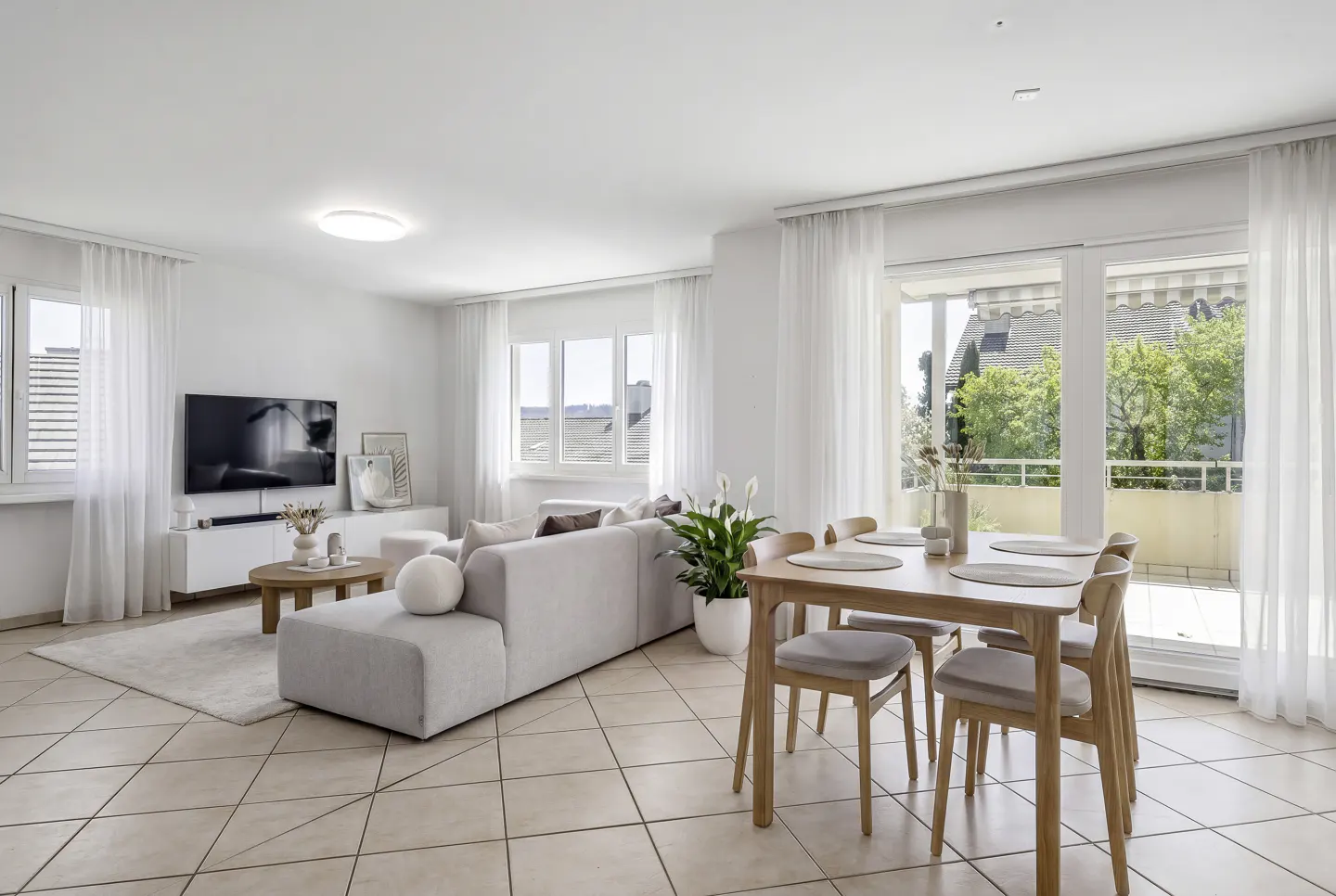 Bright, open-plan living room with a light gray sofa, wooden dining set, tiled floors, and large windows with sheer curtains.