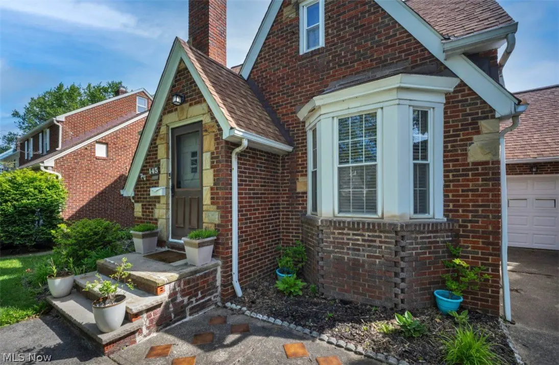 A red brick house with a brown roof, a chimney, and a small front yard with potted plants.