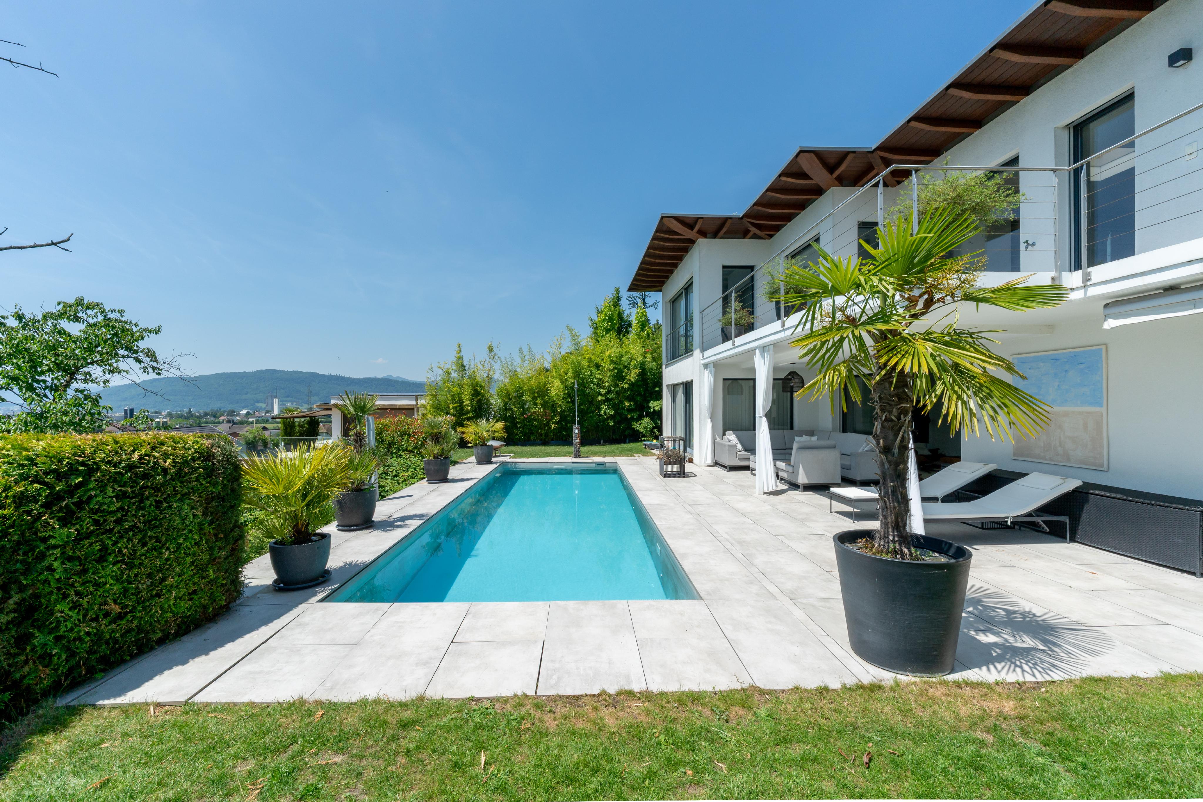 Modern white house with a rectangular pool, patio lounge chairs, potted palm trees, and a scenic mountain view.