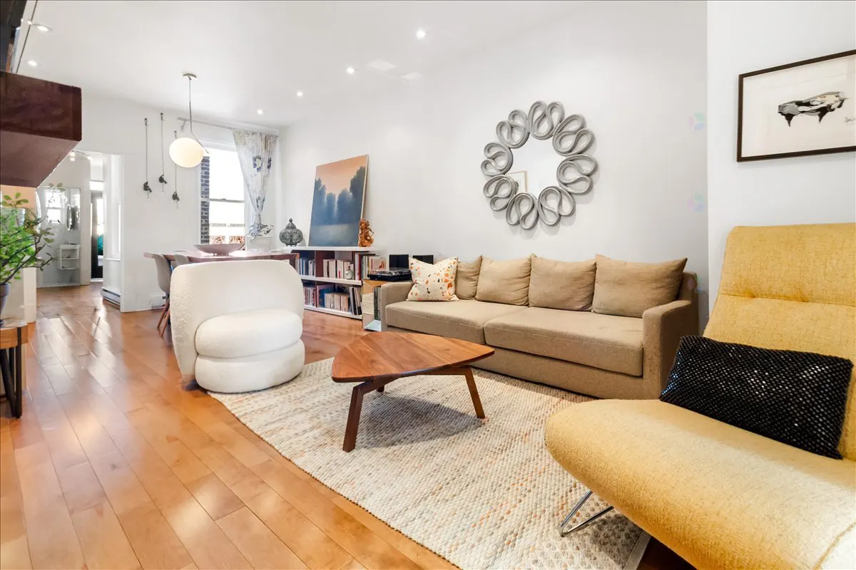 Bright living room with hardwood floors, beige sofa, yellow chair, and white accent chair. A wooden coffee table sits on a patterned rug.