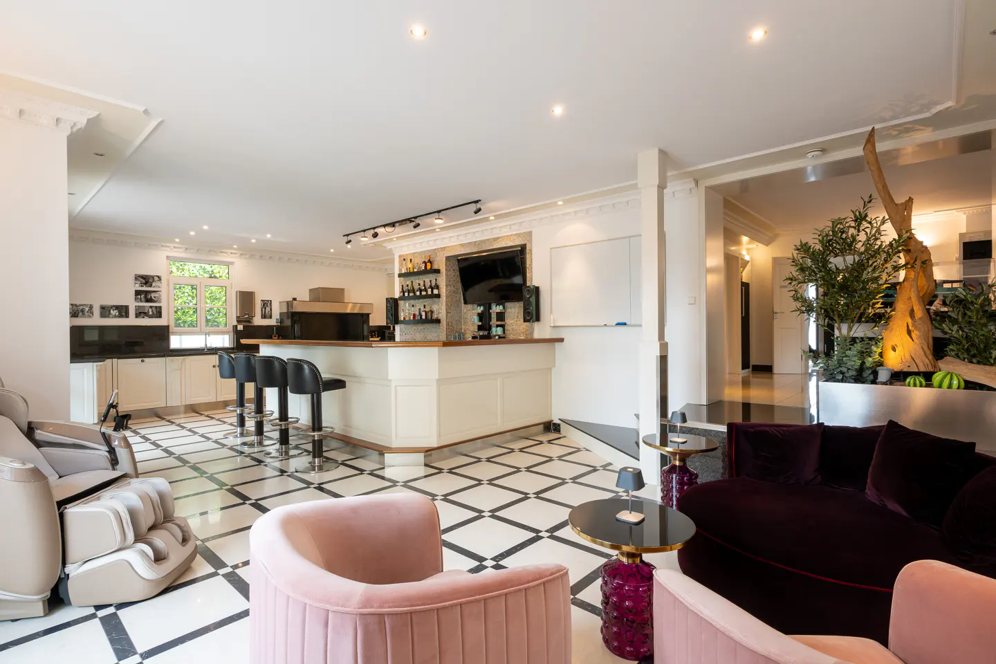 Modern open-plan living area with a bar, black stools, pink armchairs, a dark sofa, and white tiled floors with black trim.