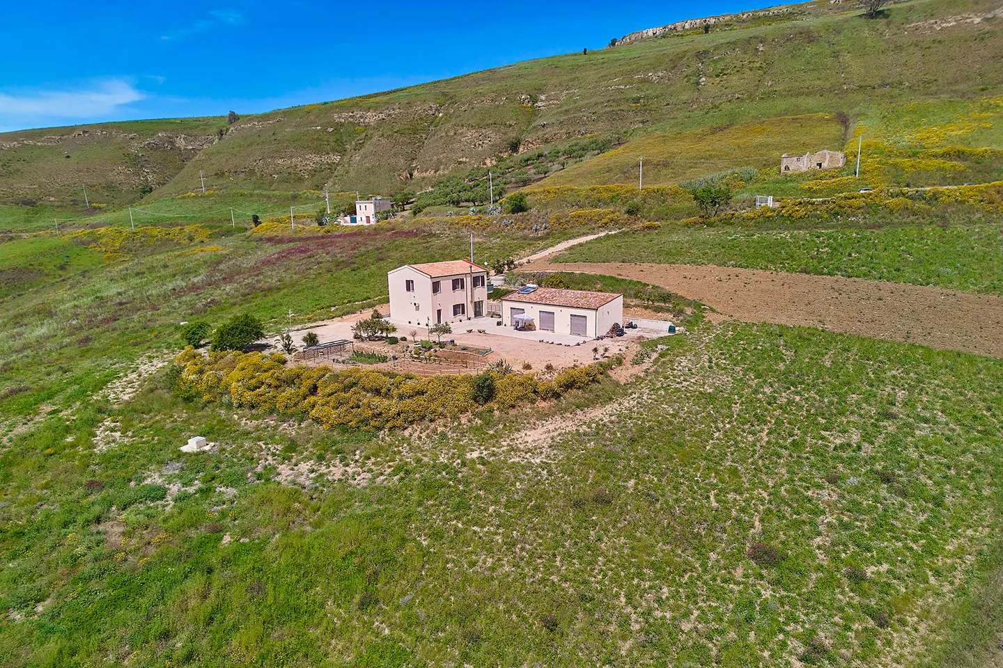 Aerial view of a beige two-story house with a red tile roof and a detached garage on a green hillside.