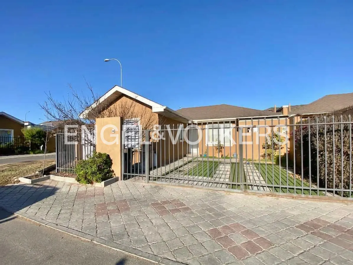 Tan single-story house with a gray metal fence and a paved driveway under a clear blue sky. "Engel & Volkers" is superimposed on the image.