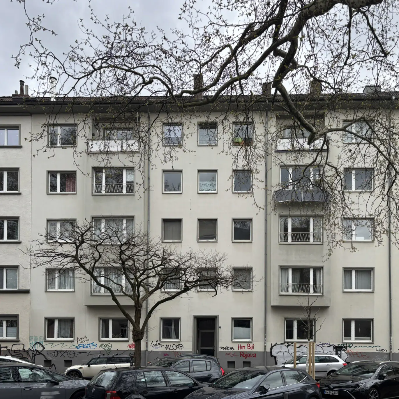 Apartment building with many windows, bare trees in front, and parked cars. The building is light gray.