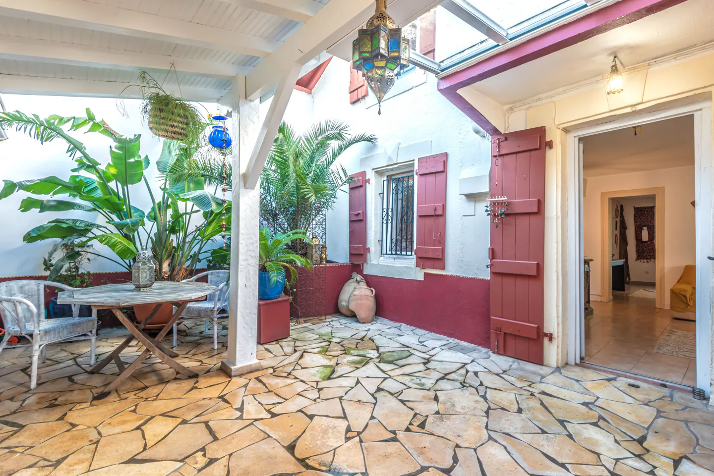 A sunlit courtyard with stone floors, a wooden table and chairs, and lush green plants. Red shutters adorn a white building with an open doorway.