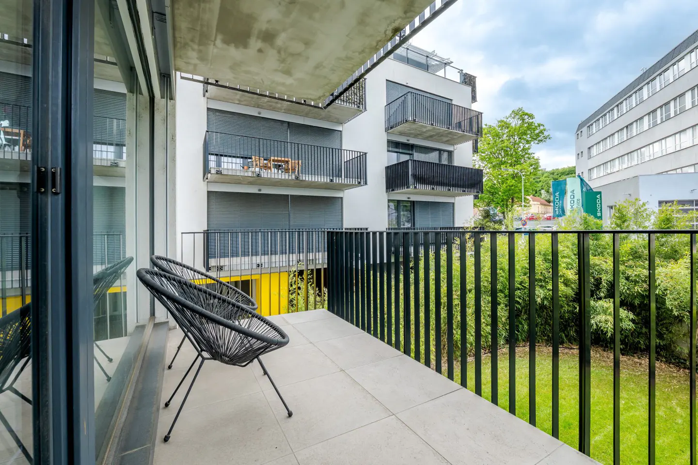Modern apartment balcony with two black woven chairs, tiled floor, and a view of a neighboring building and green garden.