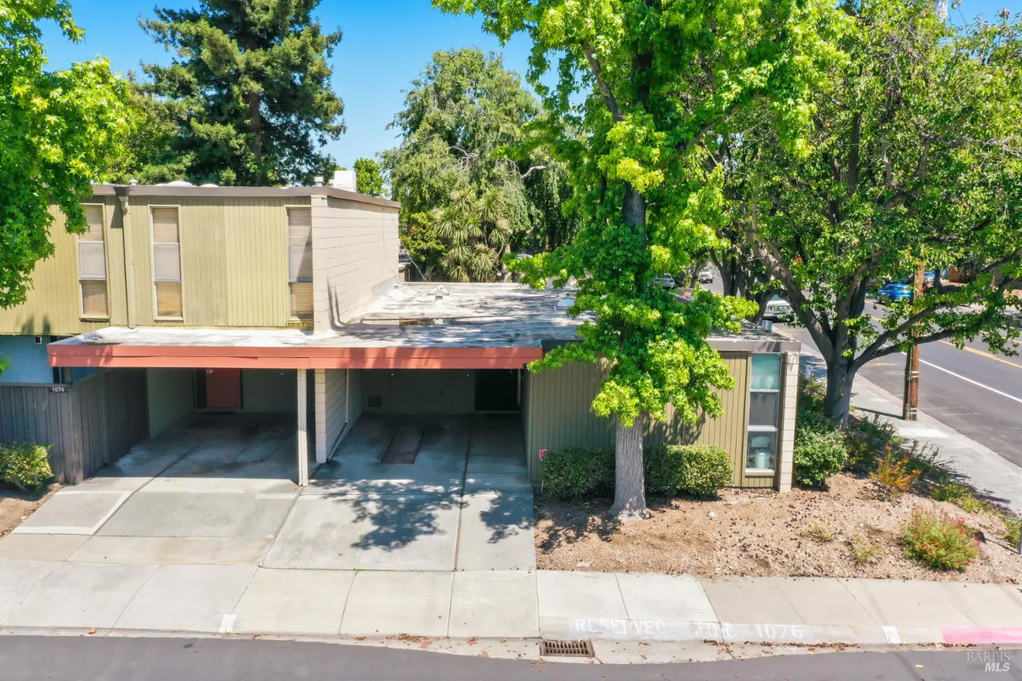 Aerial view of a two-story townhouse with a carport, olive green siding, and a flat roof. Lush green trees surround the building.