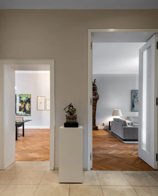 Interior view of a home with two doorways. A statue sits on a white pedestal. The rooms have wood floors and art on the walls.