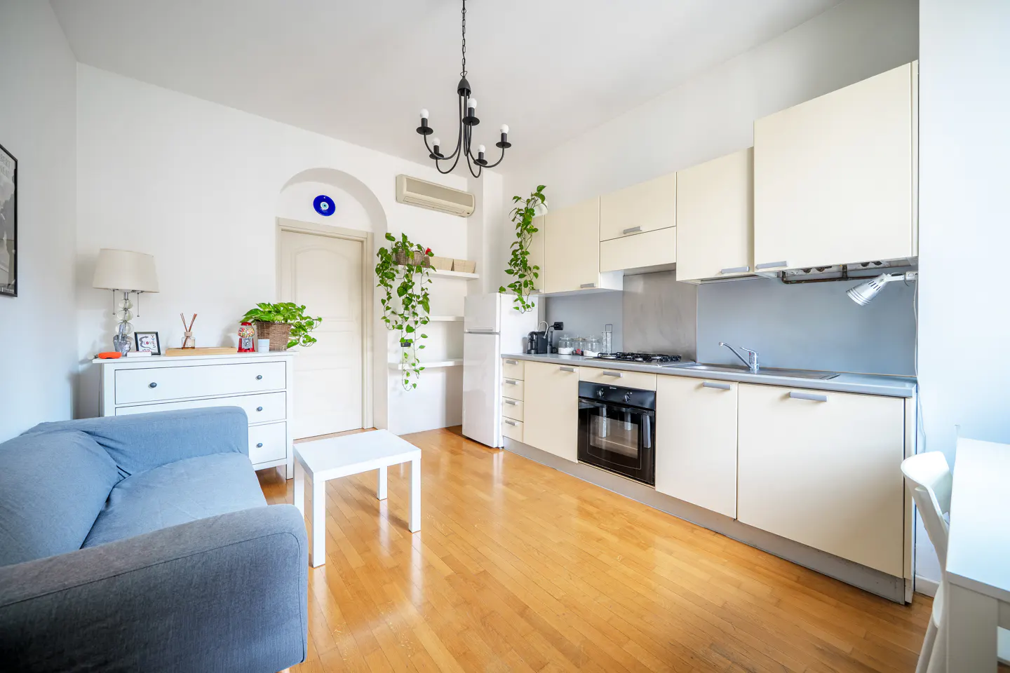 Bright, open apartment with hardwood floors. A blue sofa and white dresser are on the left, and a kitchen with cream cabinets is on the right.