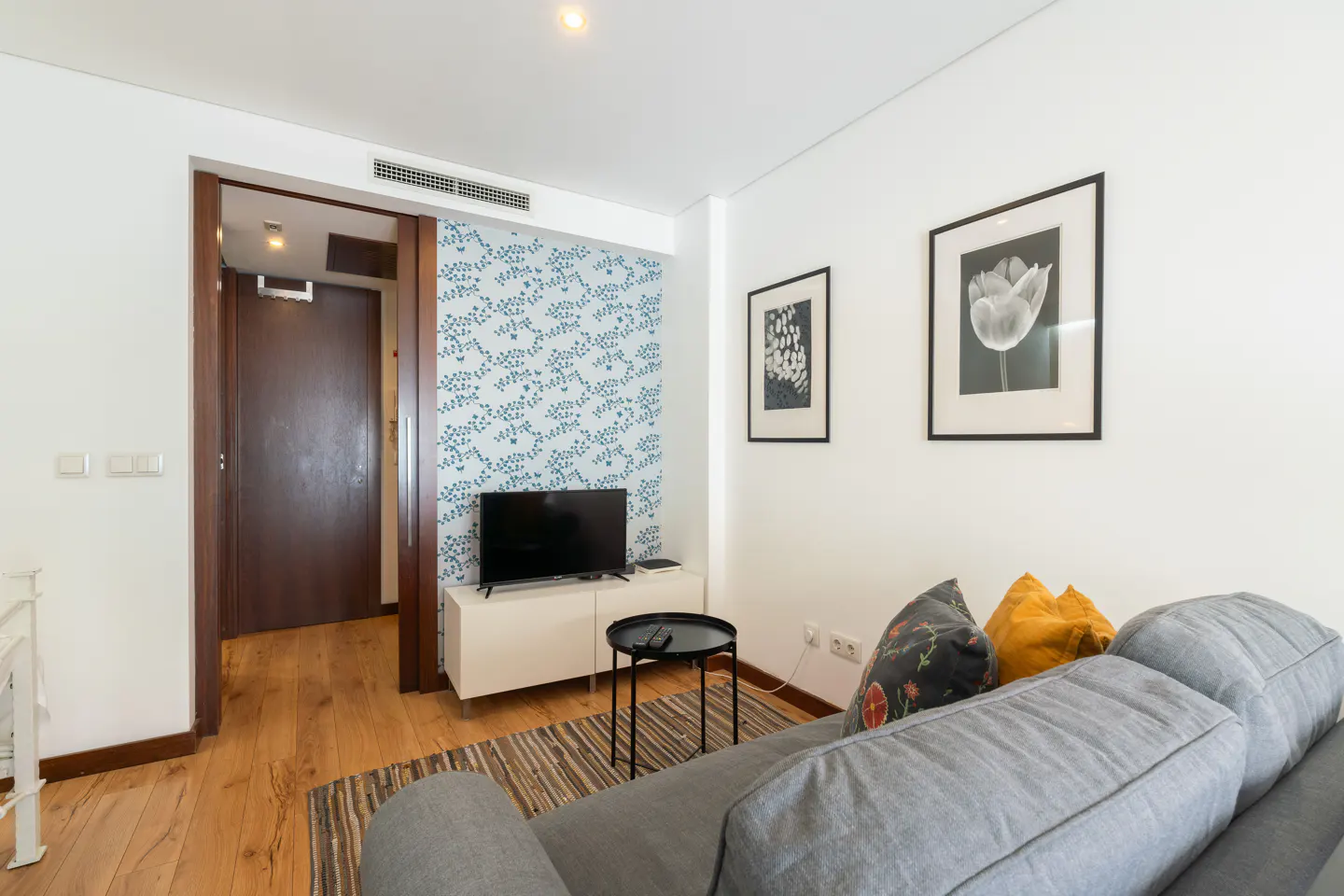 Living room with gray sofa, floral pillows, and black-framed art. A TV sits on a white cabinet against a floral wallpapered wall.
