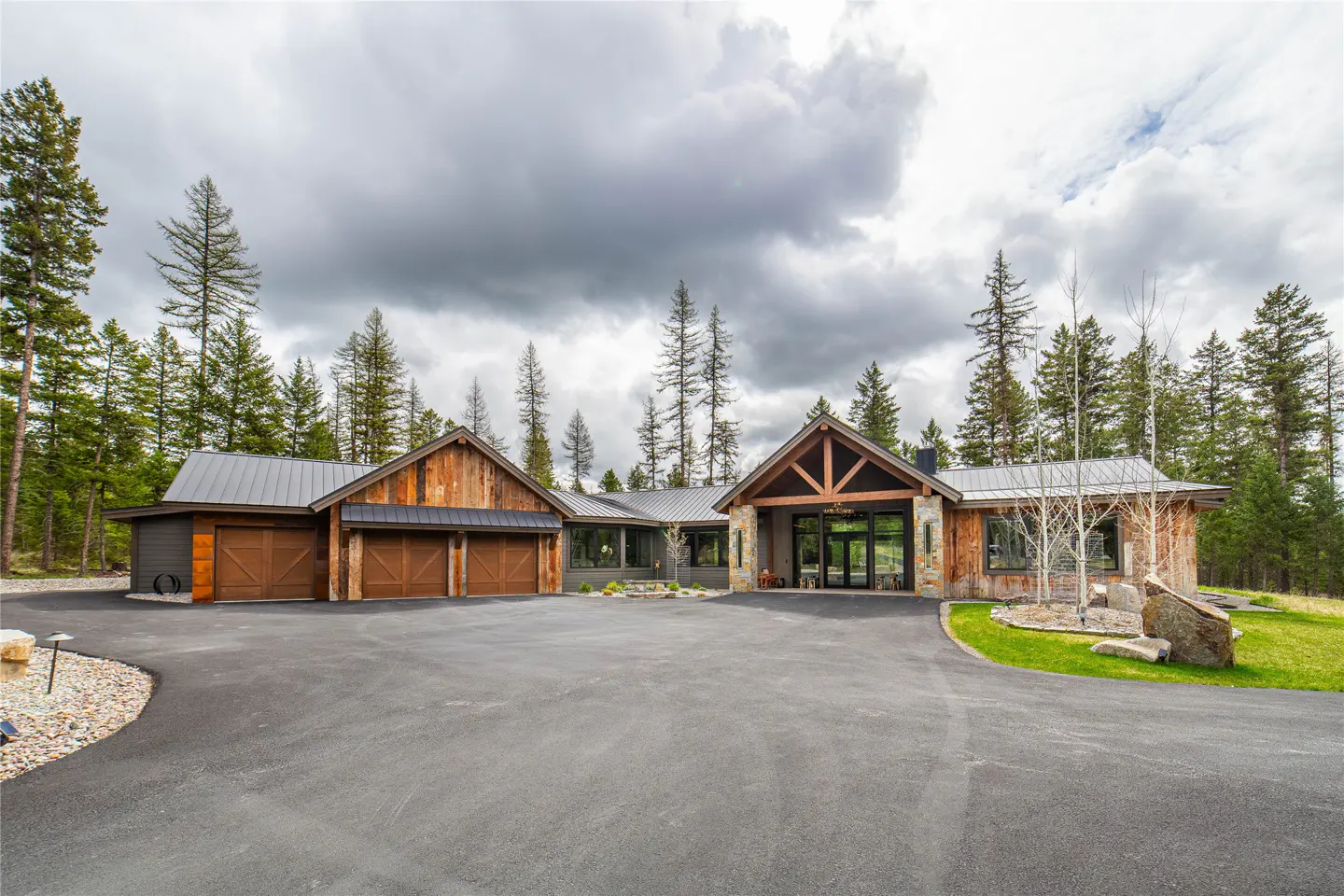 A wide shot of a rustic-style home with wood and stone accents, a metal roof, and a large driveway. Tall trees surround the property.