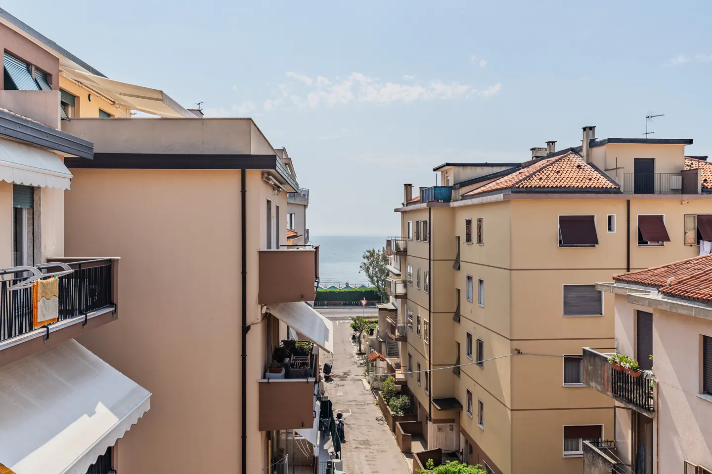 View of a street between apartment buildings leading to the ocean under a blue sky.