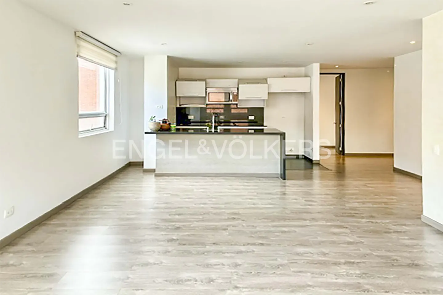 Bright, open-concept apartment with light wood floors, white walls, and a modern kitchen with a gray island. A window provides natural light.