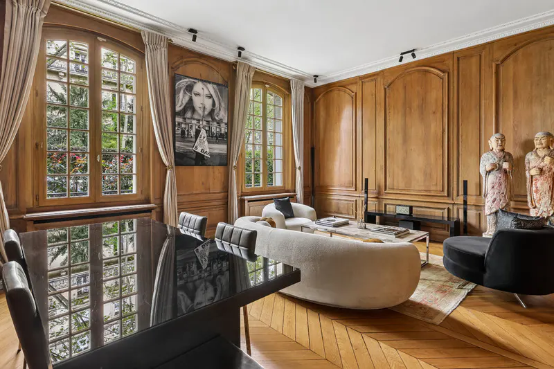 A living room with wood paneled walls, a black dining table, and a curved white sofa. Two large windows with cream curtains let in natural light.