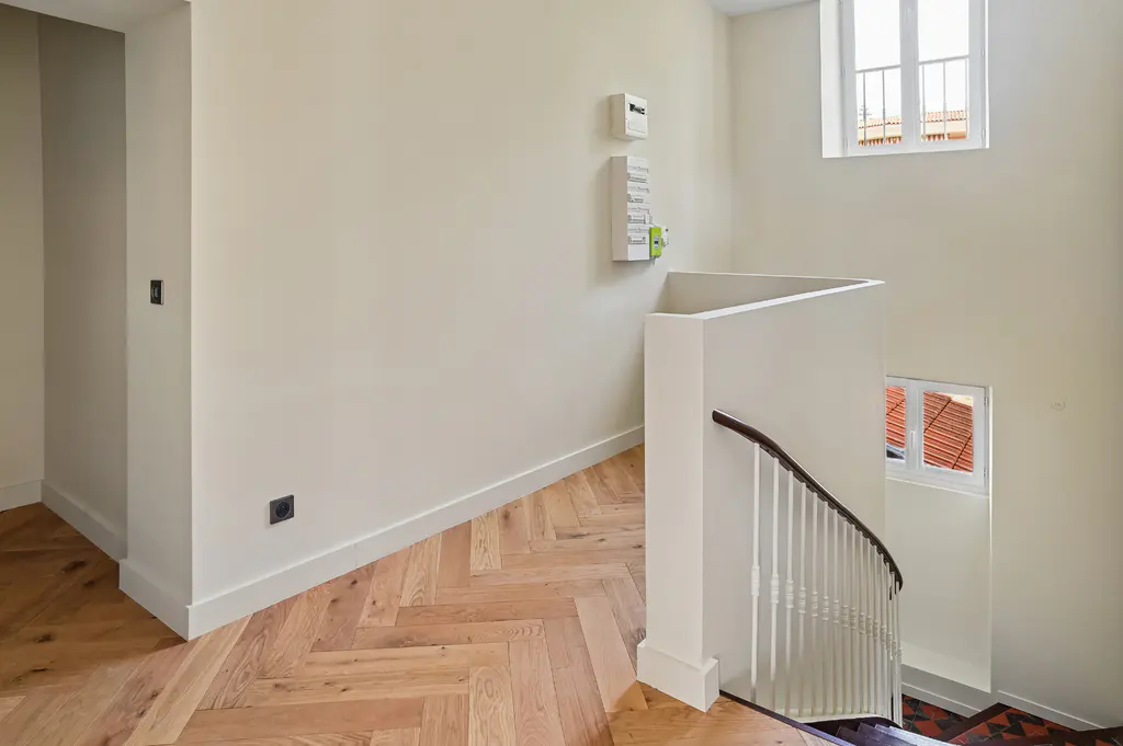 Interior view of a staircase with white walls, herringbone wood floors, and a curved railing. Windows provide natural light.