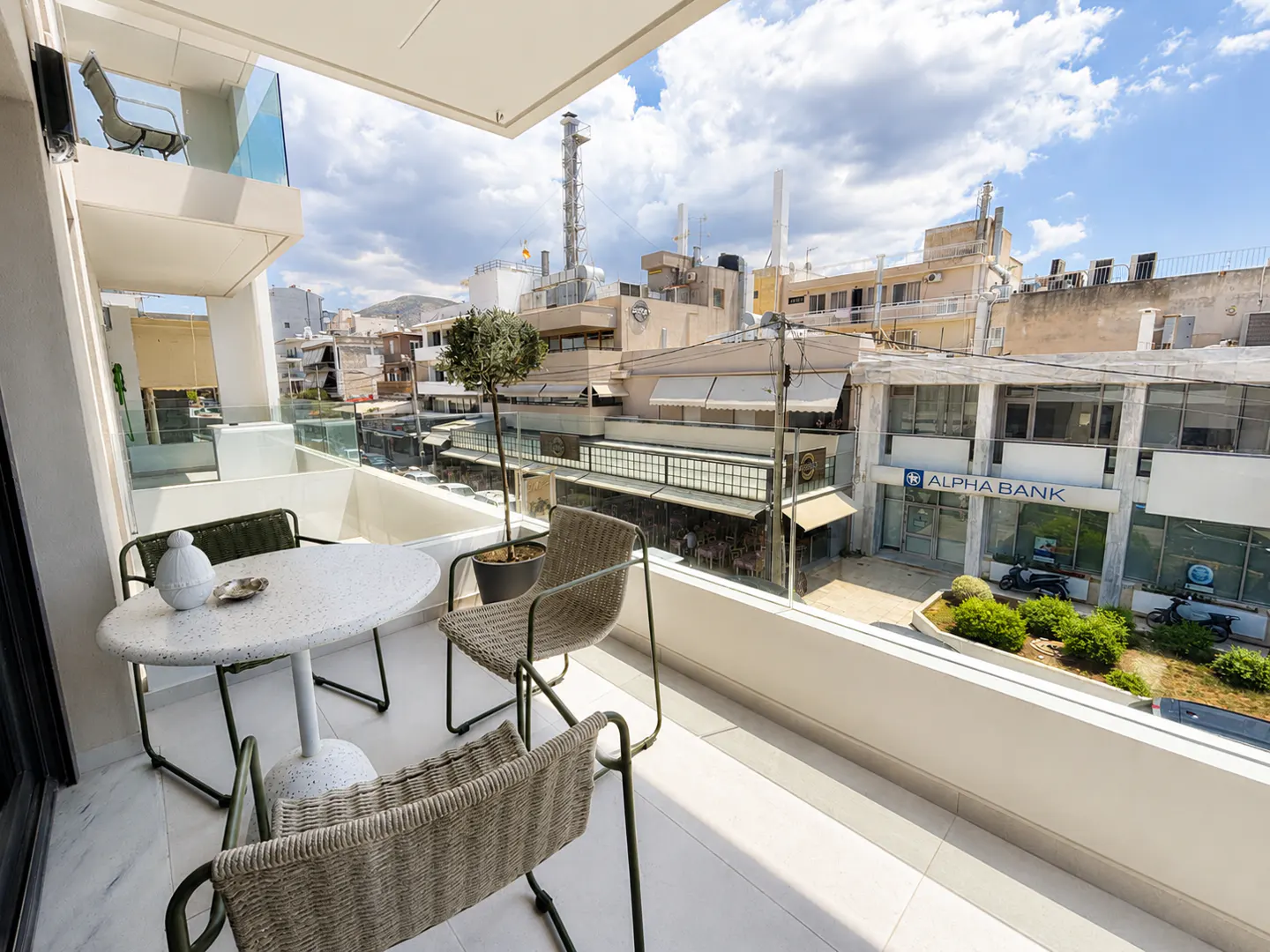 Balcony view with a round table, three chairs, and a small tree in a pot. City buildings are visible in the background.